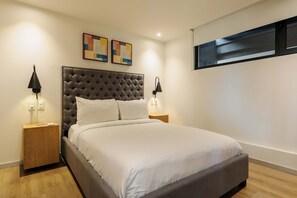 Ein Schlafzimmer mit einem gesteppten Kopfteil, weißer Bettwäsche, zwei Nachttischlampen und einem Fenster mit Jalousien.