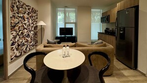 Ein modernes Wohnzimmer mit einer Couch, einem runden Tisch mit Salz- und Pfefferdosen, einem Fernseher und einer Küchenzeile inklusive Kühlschrank und Mikrowelle.