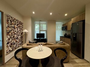 Ein modernes Wohnzimmer mit einer Couch, einem runden Tisch mit Salz- und Pfefferdosen, einem Fernseher und einer Küchenzeile inklusive Kühlschrank und Mikrowelle.