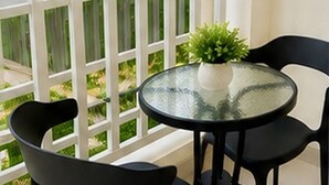 A balcony with a glass table and two chairs, a potted plant, and a white railing. - Elimstay Hospitality Jimbaran Bali | Comfort, Pool & Tropical Ambience (Badung)