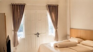 A bedroom with a bed, curtains, a door, and a television. - Elimstay Hospitality Jimbaran Bali | Comfort, Pool & Tropical Ambience (Badung)
