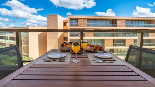 Una mesa en el balcón con alimentos para el desayuno, cielo despejado y un edificio moderno al fondo.