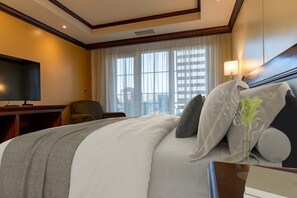 Ein Hotelzimmer mit einem großen Bett, einem Fernseher, einem Sessel und einem Fenster mit Blick auf die Stadt.
