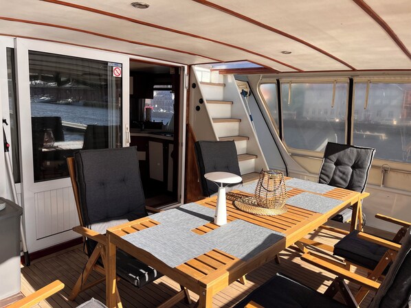 A sunlit deck with a table and chairs, a view of the water, and a staircase leading to an upper deck.