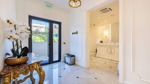 A hotel lobby with a marble floor, a gold-trimmed table with a potted plant, a door with an exit sign, and a light fixture. - Hotel Eden (Budva)
