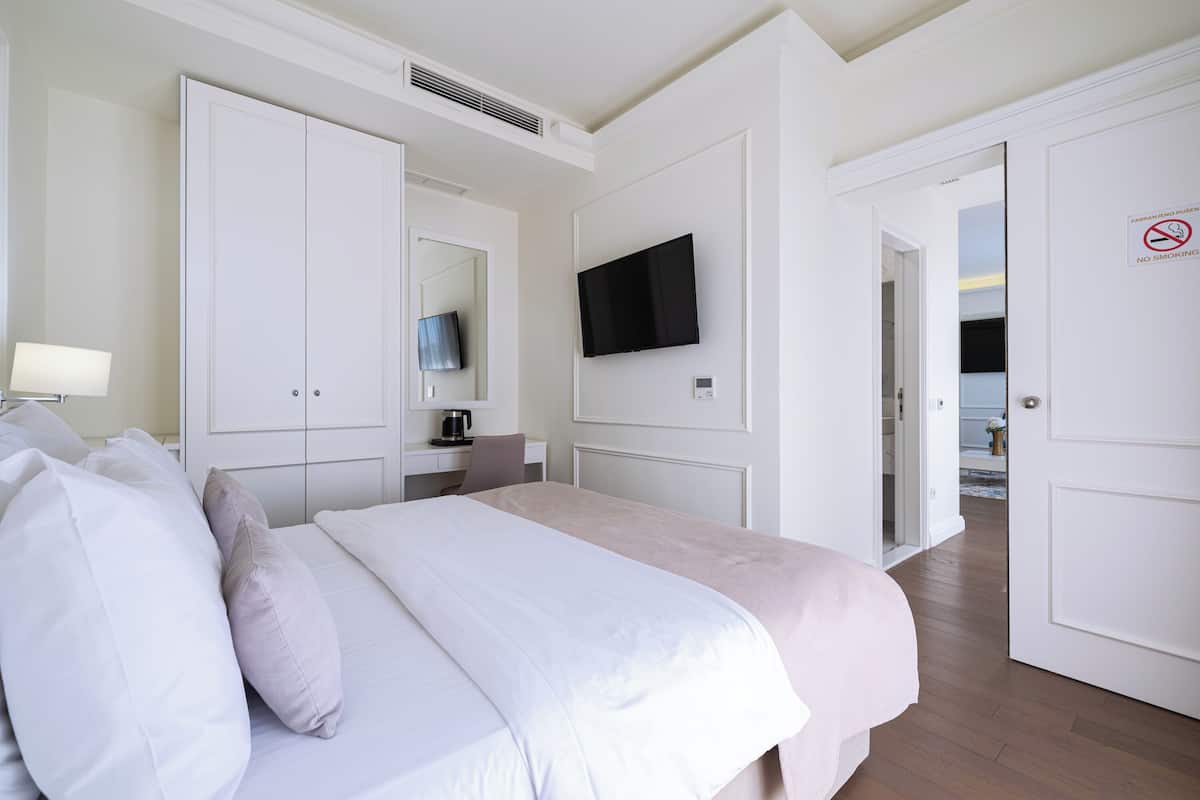 A hotel room with a bed, a TV mounted on the wall, a desk with a lamp, and a door with a no-smoking sign.