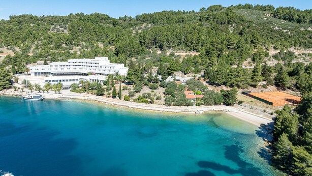 Un hotel frente al mar con un edificio grande, una playa y colinas boscosas circundantes.