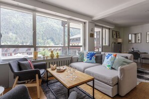 A modern living room with a sofa, armchairs, a coffee table, and a view of a forested area through large windows. - Concordia 27, Town Centre, Chamonix All Year (Chamonix)