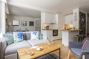 Ein modernes Wohnzimmer mit einer Couch, einem Couchtisch mit Speisen und Gläsern, einer Küchenzeile mit Schränken und Geräten sowie einem Essbereich mit Hockstühlen.