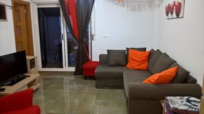 Ein Wohnzimmer mit einer Couch, roten und orangefarbenen Kissen, einem Fernseher und einer Schiebetür zu einem Außenbereich.