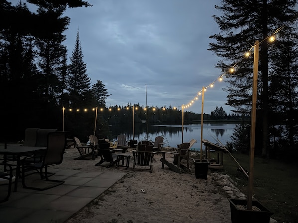 An outdoor seating area with chairs and tables, string lights, and a view of a lake surrounded by trees.