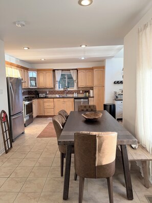 A modern kitchen with wooden cabinets, a stainless steel refrigerator, a dining table with chairs, and a window with curtains.
