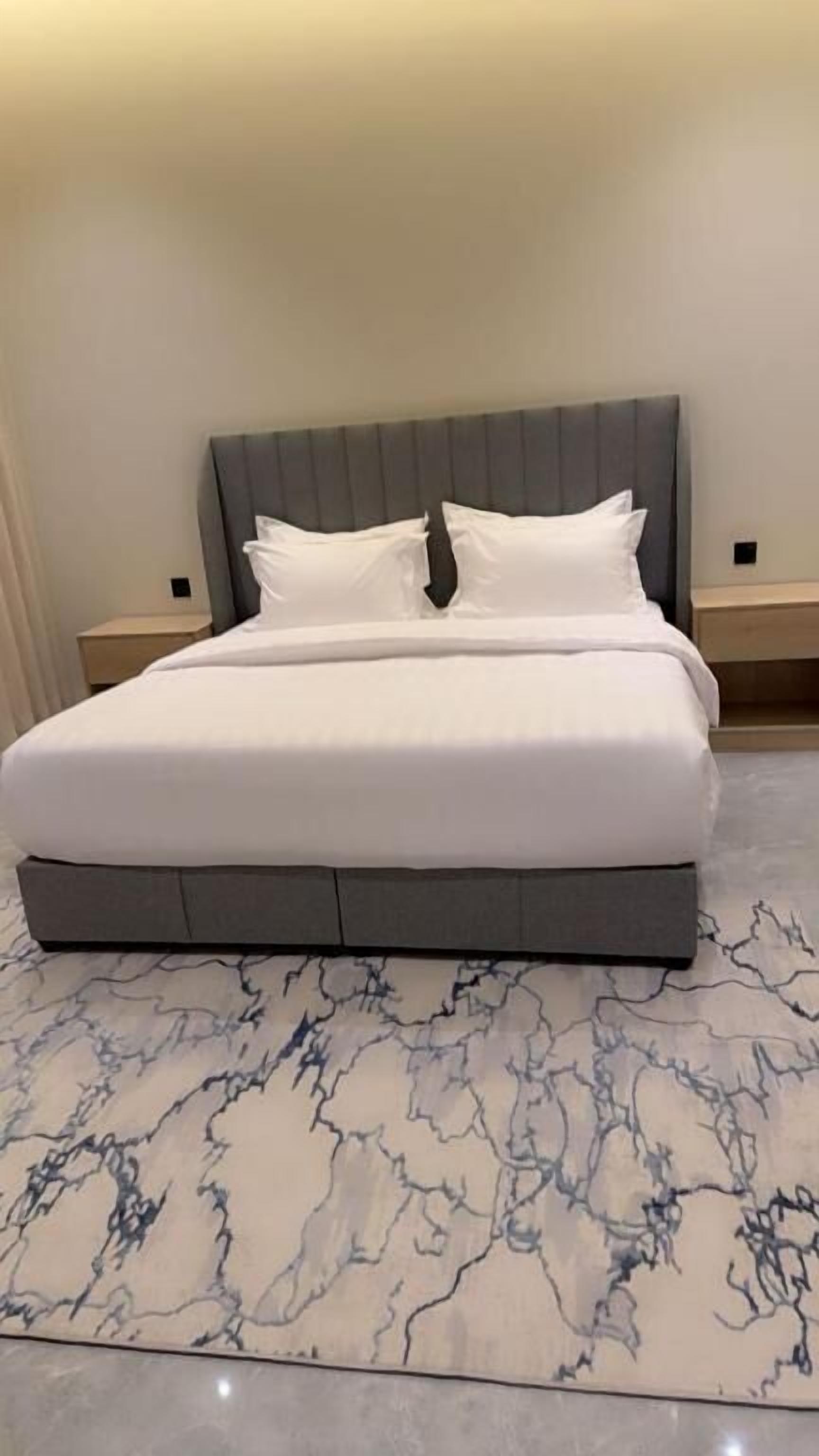 Cama com cabeceira cinza, roupa de cama branca e tapete com padrão marmorizado.