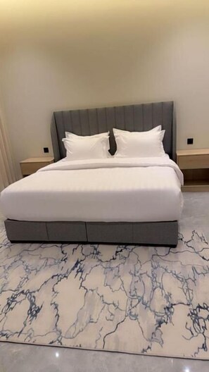A bed with a grey headboard, white bedding, and a marble-patterned rug. - Casa Residence Al Narjees (Riyadh, Al Narjis neighborhood)