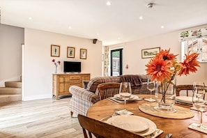 A dining area with a wooden table set for four, a sofa, and a television in the background. - Loft 1 - One bedroom house with parking (Nuneham Courtenay)