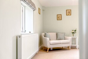 Une pièce avec un radiateur, un canapé blanc avec des coussins, une petite table sur laquelle se trouve un vase, et deux tableaux encadrés accrochés au mur.
