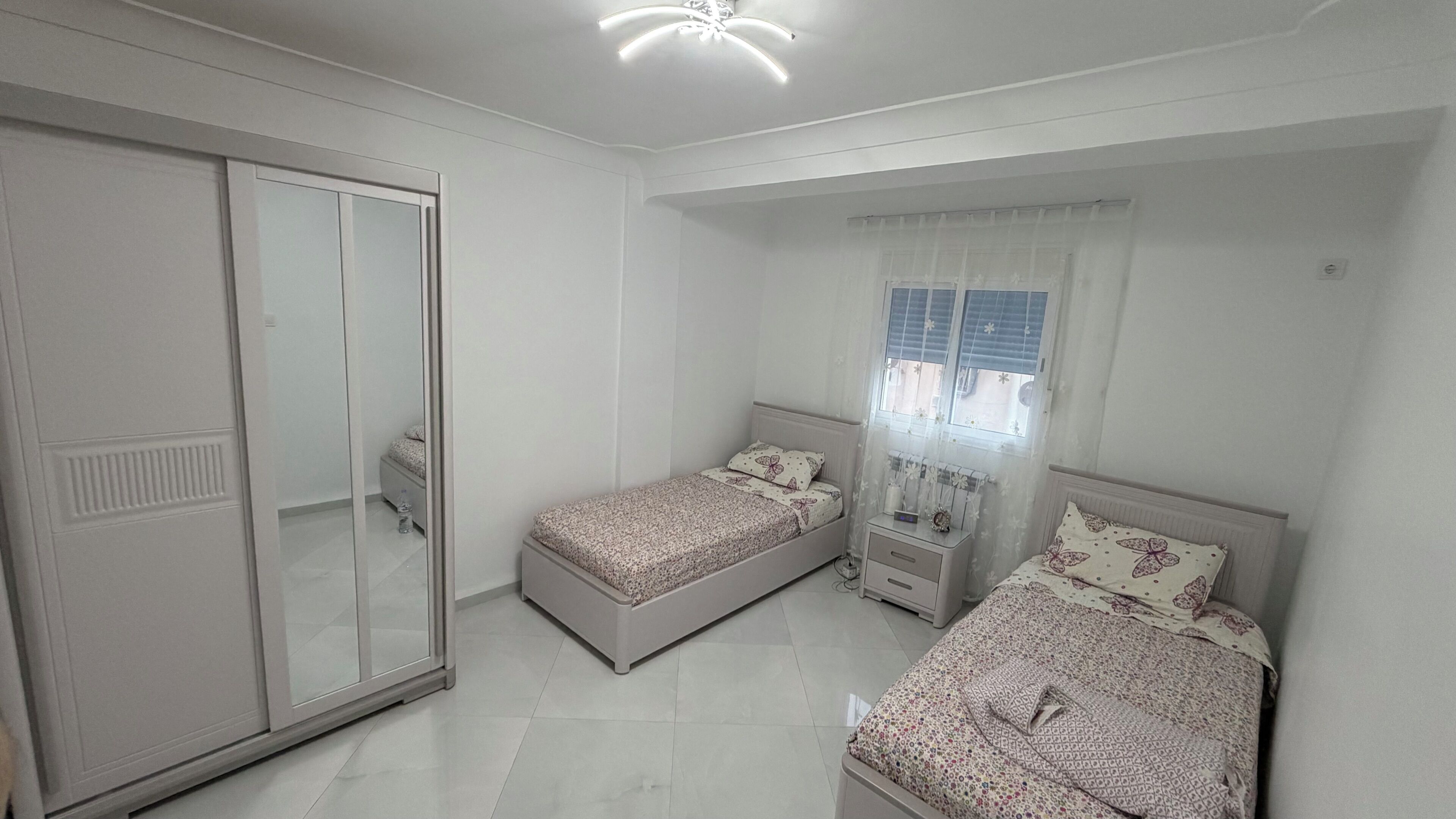 A bedroom with two single beds, a wardrobe, a nightstand, and a window with curtains.