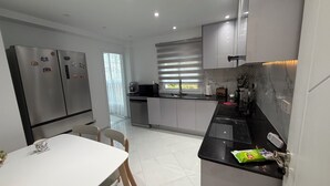 Eine moderne Küche mit Edelstahlgeräten, ein Essbereich mit einem weißen Tisch und Stühlen sowie ein Blick ins Freie durch ein Fenster mit Jalousien.