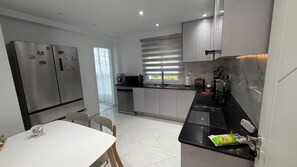 Une cuisine moderne dotée d’appareils électroménagers en acier inoxydable, un coin repas avec une table et des chaises blanches, et une vue sur l’extérieur par une fenêtre équipée de stores.