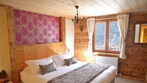 A wooden cabin bedroom with a bed, a chandelier, a window with curtains, and a patterned wall. - Chalet Cannelle (La Chapelle-d'Abondance)