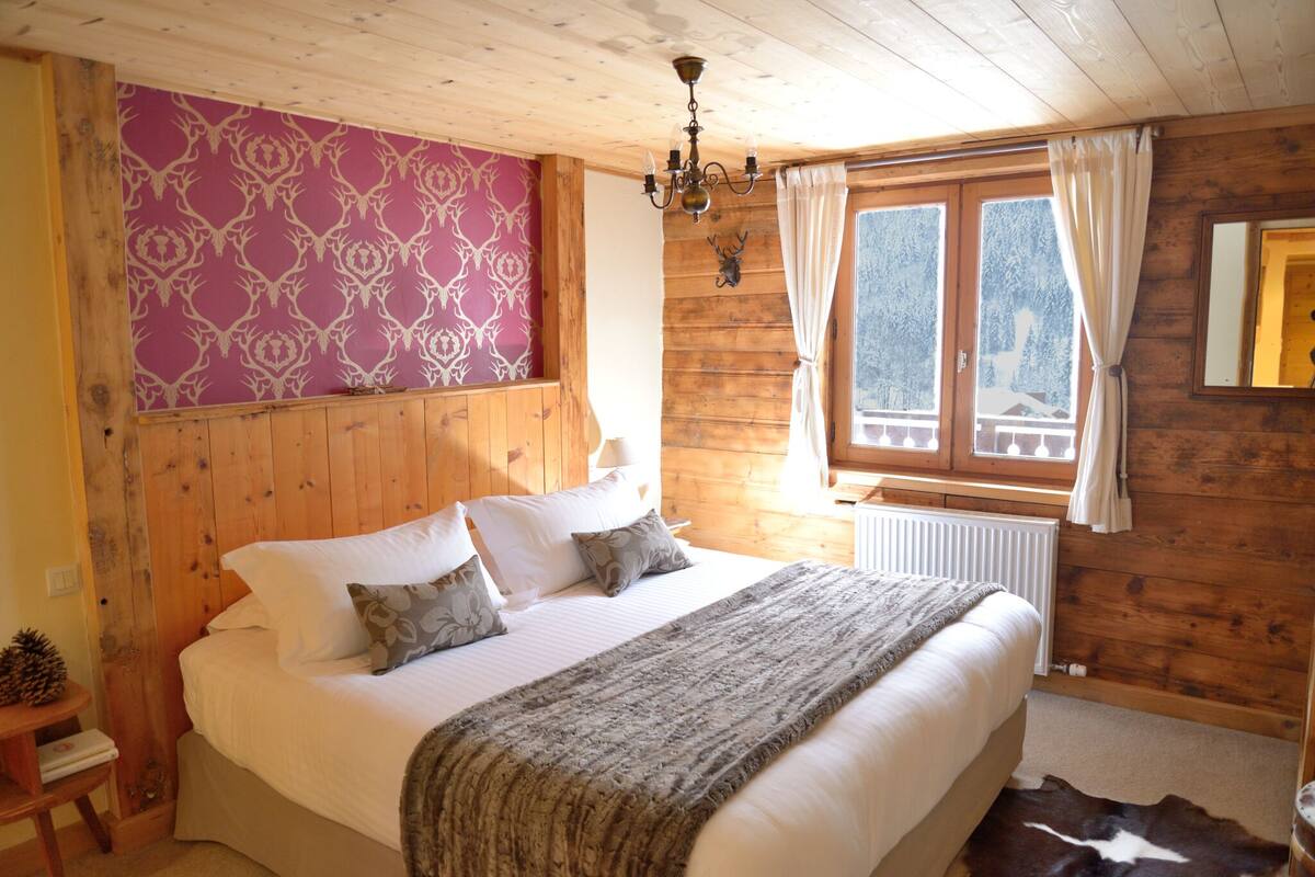 A wooden cabin bedroom with a bed, a chandelier, a window with curtains, and a patterned wall.
