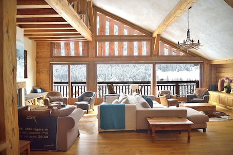 A spacious loft-style room with a high ceiling, large windows, and a balcony with a view of snow-covered trees.. Chalet Cannelle