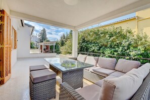 An outdoor patio with wicker furniture, a glass-top table, and a view of a house and greenery. - Villa Can Benavent by Slow Villas (Arenal)