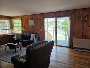 A room with wooden walls, a black leather sofa, a small table, and a large window with a view of trees and a deck. - Bridges Camp - Private Beach! Dogs welcome! (Little Deer Isle)