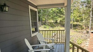 A porch with two rocking chairs and a table, a wooden railing, and a window. - Bridges Camp - Private Beach! Dogs welcome! (Little Deer Isle)