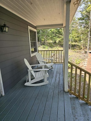 A porch with two rocking chairs and a table, a wooden railing, and a window. - Bridges Camp - Private Beach! Dogs welcome! (Little Deer Isle)