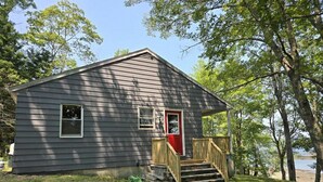 A small, single-story cabin with a front porch and a red door, surrounded by trees and a grassy yard. - Bridges Camp - Private Beach! Dogs welcome! (Little Deer Isle)