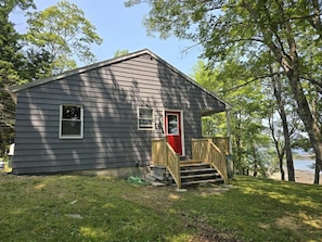 A small, single-story cabin with a front porch and a red door, surrounded by trees and a grassy yard. - Bridges Camp - Private Beach! Dogs welcome! (Little Deer Isle)