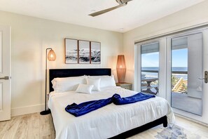 Ein Schlafzimmer mit einem Bett, einem Deckenventilator, Blick auf den Ozean durch eine Schiebetür und einem gerahmten Bild an der Wand.