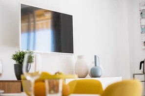 Ein moderner Essbereich mit einem weißen Tisch, gelben Stühlen, einer Topfpflanze und einem an der Wand befestigten Flachbildfernseher.