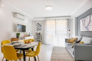 A modern living room with a grey sofa, yellow chairs, a dining table with chairs, a TV mounted on the wall, and a large window with sheer curtains. - Beautiful 2-bedroom apart. with AC in enjoyable Los Cristianos 300m to the beach (Los Cristianos)