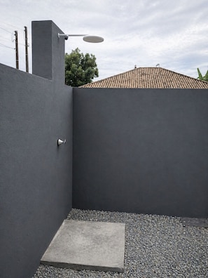A modern outdoor shower area with a concrete floor, surrounded by a dark wall and a tiled roof in the background.