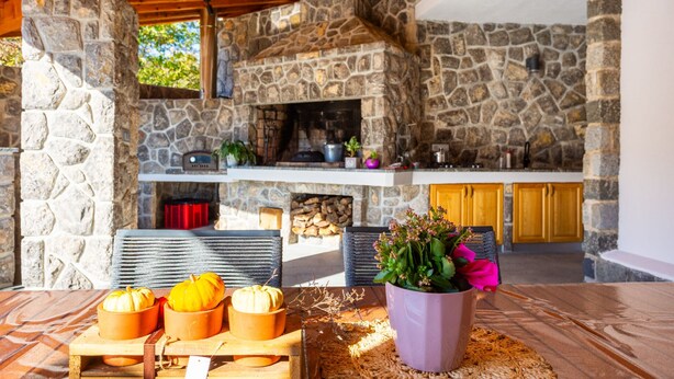 A stone fireplace with a kitchen area above it, a dining table with pumpkins, and a potted plant with pink flowers. - Apartman Mihaela with pool by Villas Guide (Kuceli)