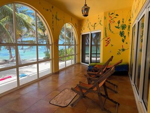 Une pièce dotée de grandes fenêtres en arc donnant sur une plage, de meubles en bois et de décorations murales aux motifs tropicaux.