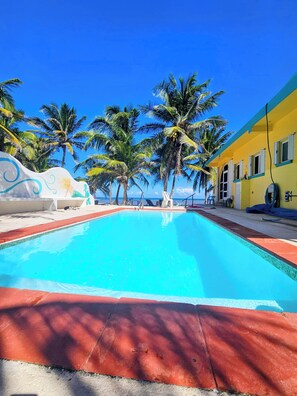 Une piscine bordée de palmiers, avec un bâtiment jaune en arrière-plan.