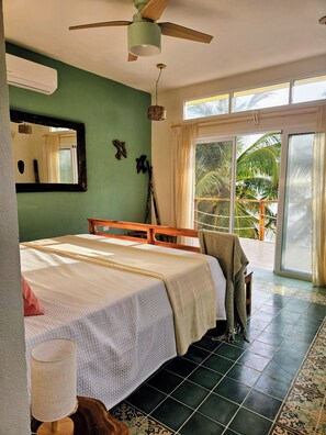 Une chambre à coucher avec un lit, un ventilateur de plafond, une fenêtre donnant sur des palmiers et un plancher carrelé.