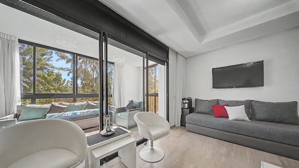 Una sala de estar moderna con un sofá gris, una silla blanca, un televisor de pantalla plana y un amplio ventanal con vistas a una zona de piscina.