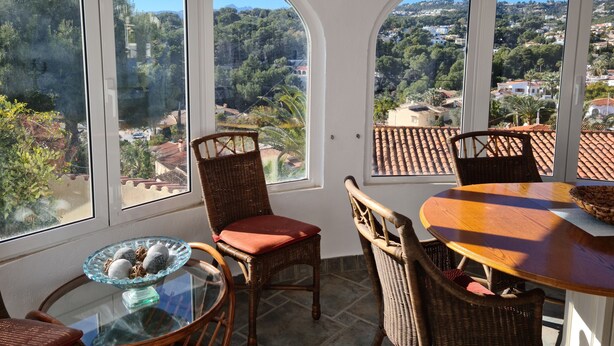 Una habitación luminosa con ventanas amplias, mobiliario de mimbre y una mesa de vidrio con adornos.