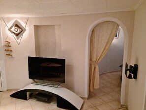 Una sala de estar con una televisión de pantalla plana sobre un soporte curvo, una mesita y un acceso a otra habitación.