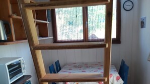 A wooden loft bed with a ladder, a kitchen area with a toaster, a dining table with chairs, and a window with blinds.