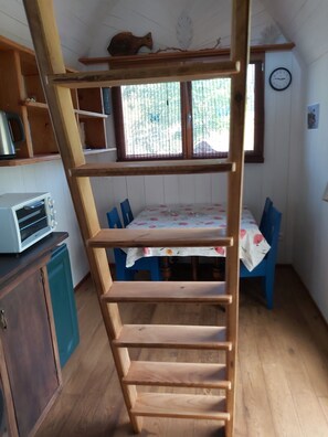 A wooden loft bed with a ladder, a kitchen area with a toaster, a dining table with chairs, and a window with blinds. - Vrbo Property (saint julien de lampon)