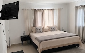 Ein Schlafzimmer mit Bett, Vorhängen, einem an der Wand montierten Fernseher und einem Nachttisch.