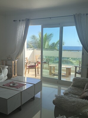 Ein modernes Wohnzimmer mit Sofa, Couchtisch und Blick auf einen Balkon mit Gartenmöbeln und Palmen.