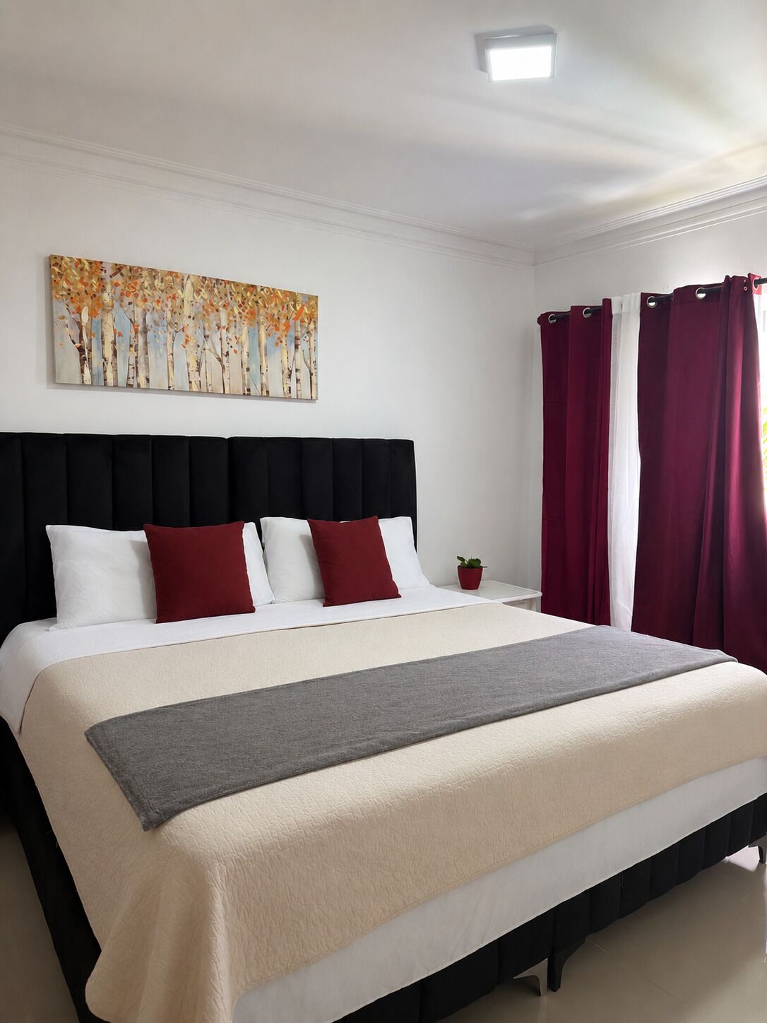 Een netjes opgemaakt bed met hoofdeinde, een nachtkastje met een plant en een schilderij aan de muur.