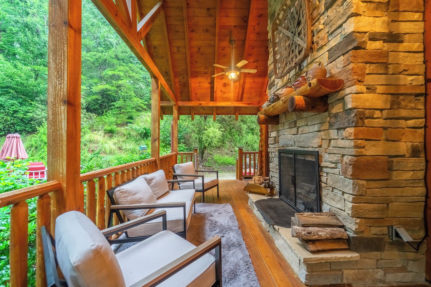 A covered outdoor seating area with a stone fireplace, wooden deck, and a view of a lush green landscape.