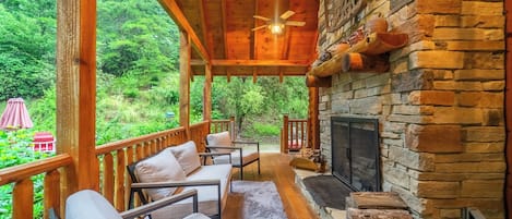 A covered outdoor seating area with a stone fireplace, wooden deck, and a view of a lush green landscape.